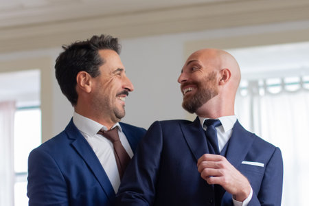 Homosexual couple in official suits smiling each other. Happy Caucasian gay couple getting dressed for wedding ceremony in hotel room, enjoying each others company. LGBT, love, marriage conceptの写真素材