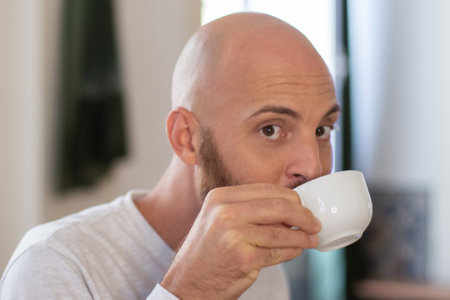 Peaceful bearded man drinking coffee at home in morning. Close-up shot of handsome Caucasian man having breakfast after waking up, sitting at table and looking at camera. Morning routine, food conceptの写真素材