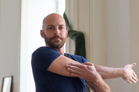 Man stretching arm at home and looking at camera. Medium shot of handsome bearded Caucasian man doing physical activity in morning, taking care of health. Sport, fitness conceptの写真素材