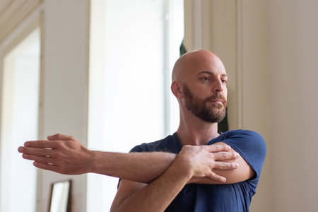 Focused man stretching arm at home in morning. Concentrated bearded Caucasian sportsman warming up before strength workout, doing physical activity after waking up. Sport, fitness conceptの写真素材