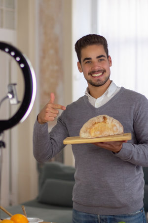 Portrait of male blogger offering healthy self-cooked bread. Smiling man in casual clothes holding wooden cutting board with loaf of bread, showing it with his finger. Vlogging about culinary conceptの写真素材