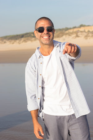 Portrait of fashionable young man on beach. Male model with shaved head in sunglasses smiling brightly, pointing at camera. Portrait, vacation, beauty conceptの写真素材