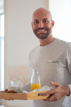 Happy homosexual man bringing his partner breakfast in bed. Vertical shot of smiling bearded man holding table tray with orange juice and buns. LGBT, food, morning routine conceptの写真素材