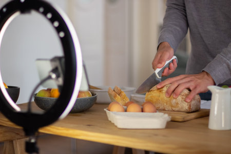 Close-up of male bloggers hands cutting loaf of bread. Young man recording live video about cooking meal at home, cutting crusty bread giving advice. Online vlogging, cooking healthy meal conceptの写真素材