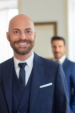 Handsome homosexual man in official suit smiling at camera. Front view of bearded man looking at camera, getting ready for wedding ceremony with partner in hotel room. LGBT, love, marriage conceptの写真素材