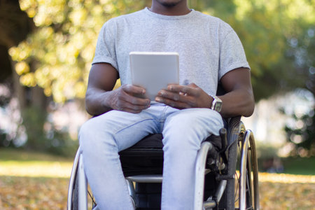 Young Black man in wheelchair spending time outside in park, holding digital tablet in his hands, relaxing on warm autumn day. Modern technologies, leisure time, disability concept.の写真素材
