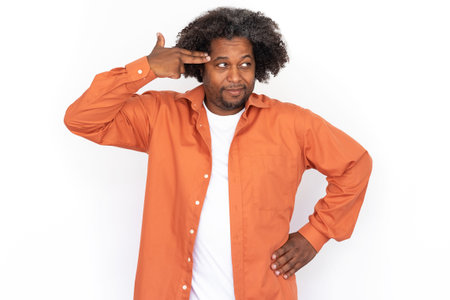 Portrait of confident mid adult man making suicide gesture against white background. African American man wearing orange shirt pointing fingers at head and looking away. Stress and boredom conceptの写真素材