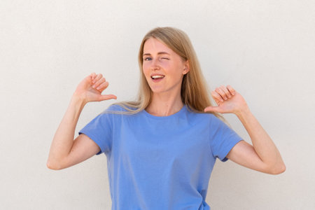 Winking young woman pointing at herself with thumbs Caucasian female model with blonde hair and blue eyes in blue T-shirt smiling and pointing at herself. Self-assurance, confidence conceptの写真素材