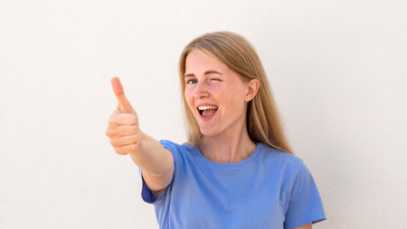 Close-up of thumb up gesture making by cheerful young woman. Caucasian female model with blonde hair and blue eyes in blue T-shirt winking and showing thumb up. Social media, advertisement conceptの写真素材