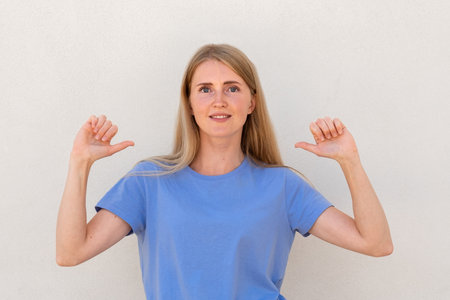 Cheerful young woman pointing at herself with thumbs. Caucasian female model with blonde hair and blue eyes in casual clothes smiling and pointing at herself. Self-assurance, confidence conceptの写真素材