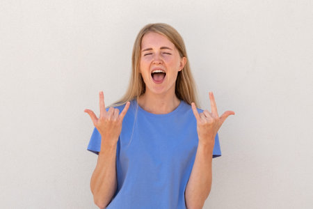 Emotive young woman showing rock-n-roll gesture. Caucasian female model with blonde hair and blue eyes in casual clothes showing horns up and screaming. Lifestyle, rebellion conceptの写真素材