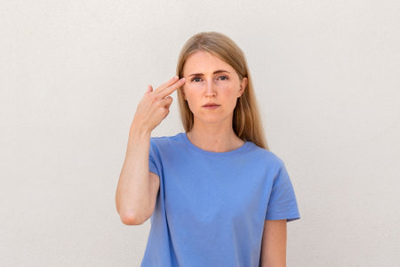 Bored young woman making suicide gesture. Caucasian female model with blonde hair and blue eyes in blue T-shirt pointing hand and fingers to head like gun. Emotions, boredom conceptの写真素材