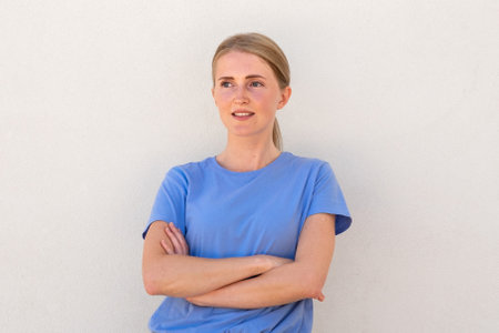 Confident young woman with crossed arms looking away. Caucasian female model with blonde hair and blue eyes in blue T-shirt crossing her arms and watching something. Self-assurance concept.の写真素材