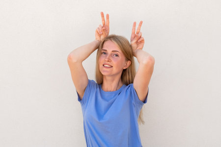 Cheerful young woman showing bunny ears with her fingers. Caucasian female model with blonde hair and blue eyes in blue T-shirt holding victory sign above head. Fun, entertainment conceptの写真素材