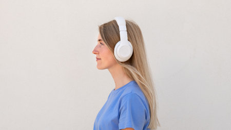 Relaxed young woman listening to music in headphones standing aside. Caucasian female model with blonde hair and blue eyes in casual clothes enjoying music. Lifestyle, music conceptの写真素材