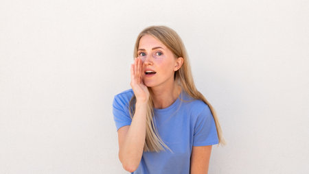 Close-up of whispering young woman holding hand near mouth. Caucasian female model with blonde hair and blue eyes in blue T-shirt telling something confidential. Secrecy, privacy concept.の写真素材