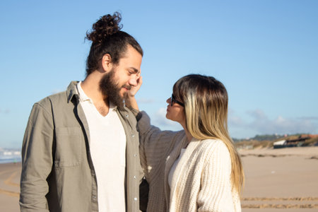 Loving couple walking on beach. Bearded man and woman in glasses looking at each other, girlfriend touching boyfriends cheek tenderly. Love, vacation, affection conceptの写真素材