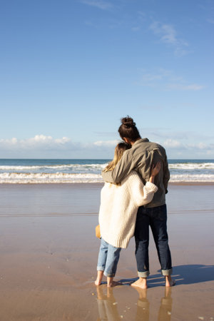 Romantic couple spending time at the beach on sunny day. Tall guy with man bun and blonde lady in cardigan standing on wet sand and hugging while enjoying picturesque sea view. Romance, travel conceptの写真素材