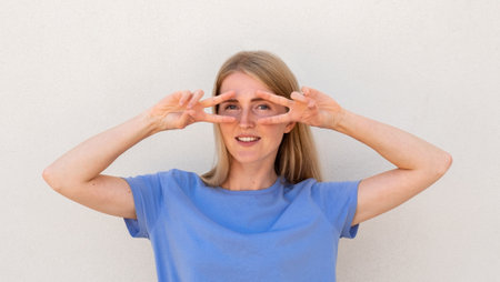 Cheerful young woman showing V-signs near her eyes. Caucasian female model with blonde hair and blue eyes in blue T-shirt smiling and holding victory signs near eyes. Posing, entertainment conceptの写真素材
