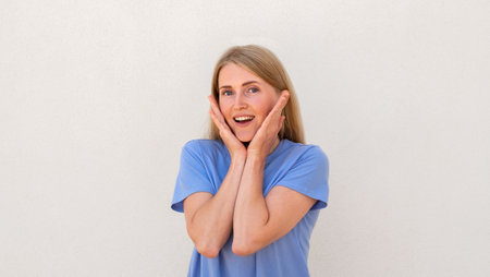 Amazed young woman holding her face with hands Caucasian female model with blonde hair and blue eyes in blue T-shirt smiling and opening her mouth from surprise. Emotions, astonishment conceptの写真素材