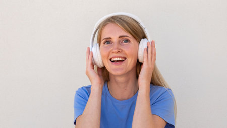Close-up of ecstatic young woman listening to music in headphones. Caucasian female model with blonde hair and blue eyes in blue T-shirt smiling and enjoying music. Lifestyle, music conceptの写真素材