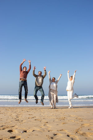 Happy senior friends having fun at beach on sunny autumn day. Four people laughing and jumping up...