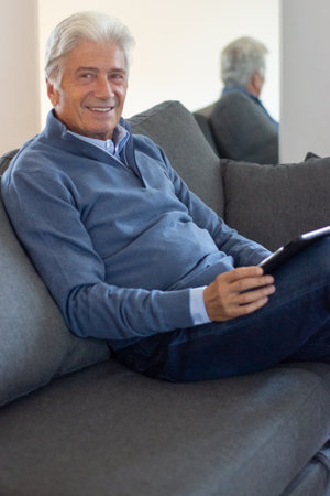 Portrait of smiling senior man with tablet. Positive aged man with gray hair resting in living room. Retirement conceptの写真素材