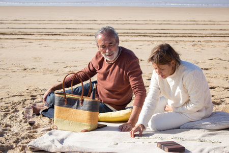 Senior couple playing dominoes while having picnic at the beach on sunny autumn day. Gray-haired man...