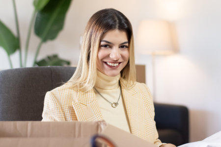 Portrait of smiling young Hispanic businesswoman in yellow checkered jacket sitting in cozy office. Intern working at post office. Delivery service conceptの写真素材
