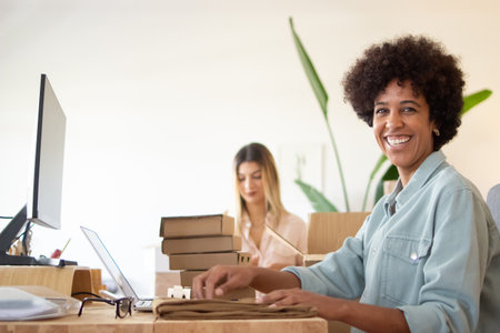 Portrait of cheerful black woman in storeroom. Laughing employee packing garment for dispatche and sitting at wooden table with computers. Online shop conceptの写真素材