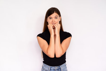 Portrait of worried young woman biting nails over white background. Caucasian lady wearing black...