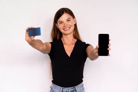 Portrait of happy young woman showing smartphone and credit card. Caucasian lady wearing black...