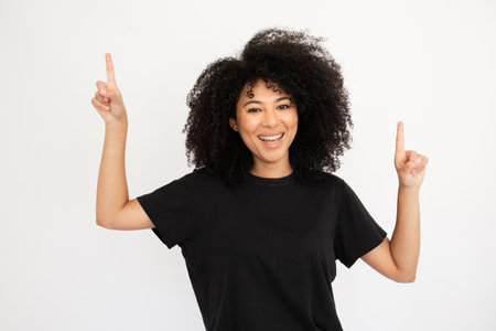 Cheerful young woman pointing above at copyspace. Hispanic female model with afro hairstyle and brown eyes in casual clothes presenting advertisement. Advertising, social media conceptの写真素材