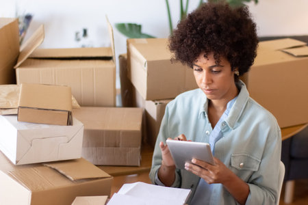 Busy African American woman using tablet in office full of cardboard boxes. Postal employee working with online order. Online startup conceptの写真素材