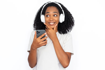 Portrait of pensive young woman with phone listening to music in headphones. African American lady...
