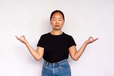 Portrait of happy young woman meditating with closed eyes over white background. Asian lady wearing black T-shirt and jeans practicing yoga. Harmony and balance conceptの写真素材