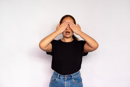Portrait of young woman covering eyes with hands over white background. Asian lady wearing black T-shirt and jeans waiting for surprise. Anticipation and surprise conceptの写真素材