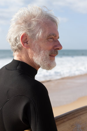 Portrait of senior man with surfboard near ocean. Thoughtful grey-haired man in wetsuit and with...