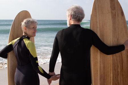 Back view of aged couple ready for surfing in cool sea. Gray-haired man and woman in wetsuits standing with boards holding hands to start riding. Relations and active lifestyle of aged people conceptの写真素材