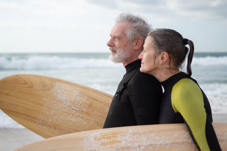 Side view of happy aged couple surfing together. Sporty man and woman in wetsuits standing on sea shore with surfboards, hugging, dreamily looking far. Sport activity, relations of aged people conceptの写真素材