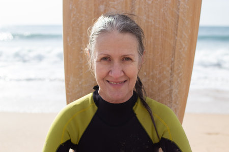 Portrait of happy senior female surfer on sea shore. Smiling Caucasian woman in wetsuit enjoying day...
