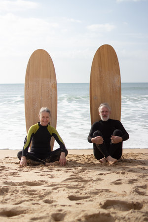 Portrait of aged couple sitting on shore with surfboards. Smiling man and woman resting on sand with boards standing behind them and looking at camera. Active rest outdoors for mature people conceptの写真素材