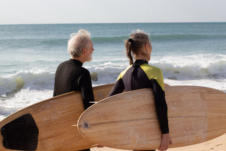 Back view of aged couple carrying big surfboards. Grey-haired man and woman in wetsuits walking along sea shore before starting surfing. Sport activity and healthy lifestyle of aged people conceptの写真素材
