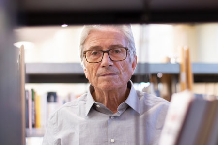 Portrait of thoughtful aged man. Gray-haired Caucasian man standing near bookshelves in public library choosing books about new technologies and looking at camera. Studying for adult people conceptの写真素材