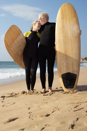 Portrait of aged male and female surfers hugging on shore. Happy man and woman standing close with surfboards embracing and man kissing womans face. Relations, healthy lifestyle of aged people conceptの写真素材