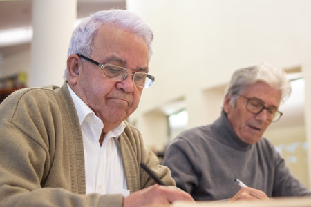 Serious aged men studying in library. Two gray-haired men in glasses sitting together at table reading about modern IT programs, making notes in notebooks. Adult people education conceptの写真素材
