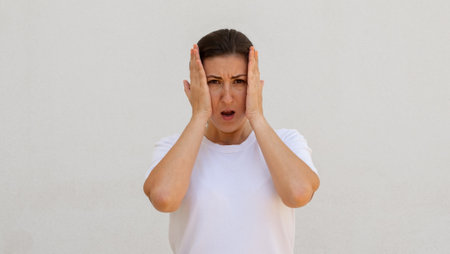Portrait of frustrated young woman holding face in hands over white background. Caucasian woman...