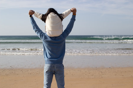 Back view of Japanese family walking on beach together. Daughter riding on fathers back on sunny day, looking at water. Leisure, vacation, parenting conceptの写真素材