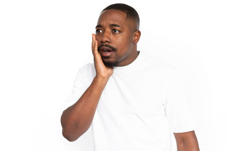 Worried African American man touching face. Portrait of shocked young male model with short hair in white T-shirt looking away with palm on cheek and open mouth. Shock conceptの写真素材