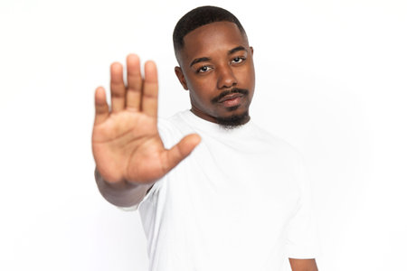 Serious African American man with open palm. Portrait of strict young male model with short hair and beard in white T-shirt looking at camera, showing stop sign. Restriction, refusal conceptの写真素材